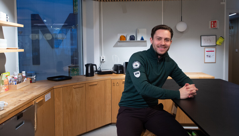 Ung mann sitter ved et høyt bord i et moderne kontorkjøkken med treverk og spotbelysning.