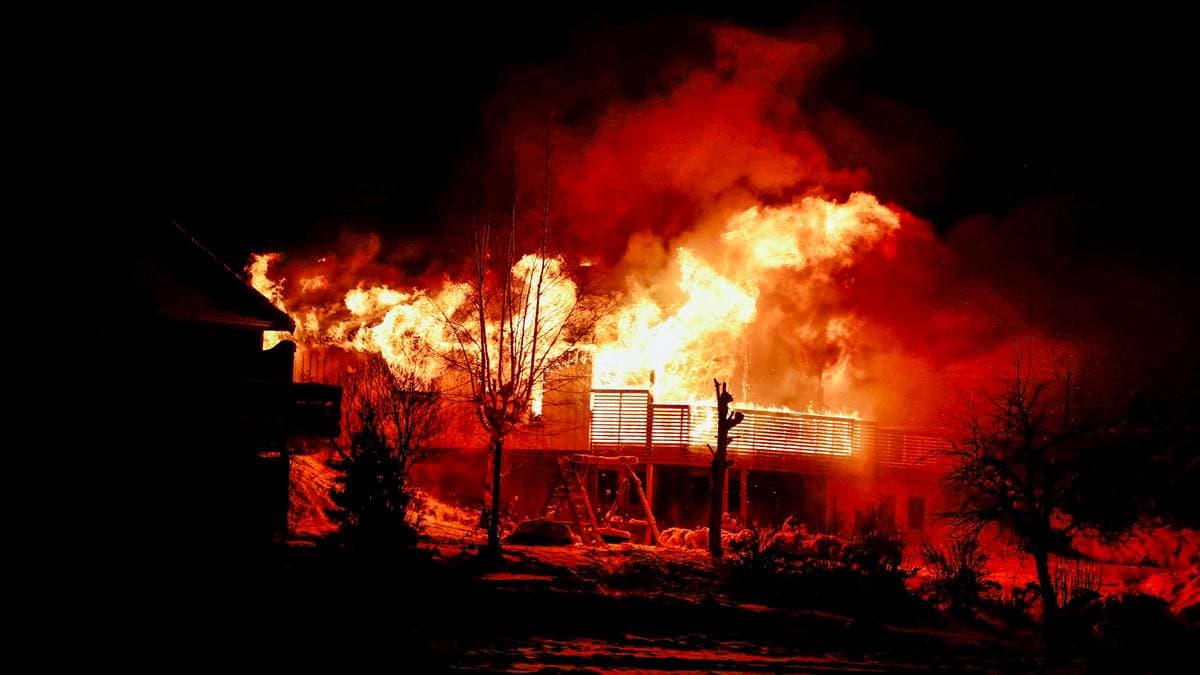 En bygning er i brann, med flammer som raser opp mot nattehimmelen. Omgivelsene er mørke, med trær og en annen bygning synlig i forgrunnen. Lysene fra flammene belyser området rundt. Det er ikke synlige personer i bildet. (Bildebeskrivelsen er laget av en KI-tjeneste)