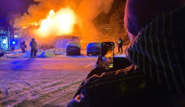 En stor brann raser i en bygning, som er omringet av flammer og svart røyk. Brannmannskap arbeider med slanger fra en brannbil som står parkert i nærheten. En person står i forkant av bildet og filmer hendelsen med en mobiltelefon. Det er snø på bakken, og kveldens mørke gir en dramatisk kontrast til brannens lys.