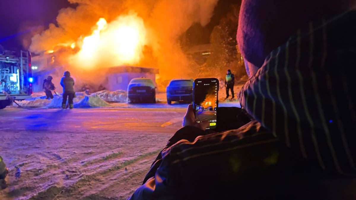 En stor brann raser i en bygning, som er omringet av flammer og svart røyk. Brannmannskap arbeider med slanger fra en brannbil som står parkert i nærheten. En person står i forkant av bildet og filmer hendelsen med en mobiltelefon. Det er snø på bakken, og kveldens mørke gir en dramatisk kontrast til brannens lys.