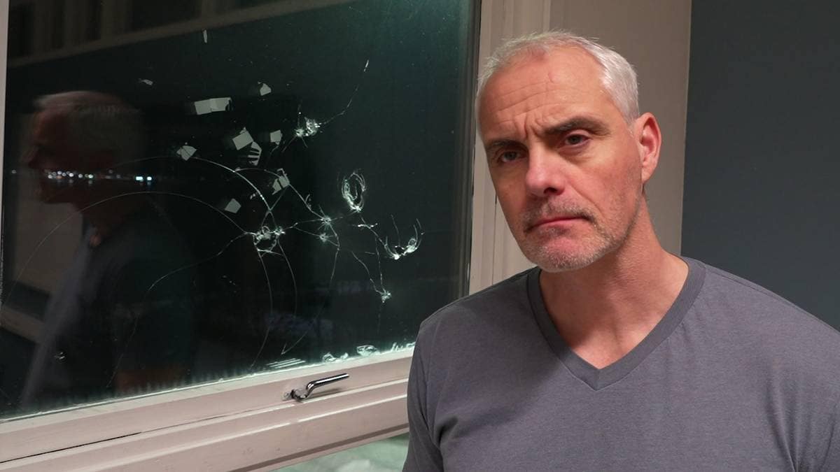 Bildet viser en mann iført en grå T-skjorte, som står foran et vindu med skader. Vinduet har sprukket glass med flere brudd og merker på overflaten. Bakgrunnen viser en mørk værelse, delvis opplyst av kjørelys utenfor. Mannen ser ut til å være i fokus, men ansiktet er uklart. Beskrivelsen er laget av KI.