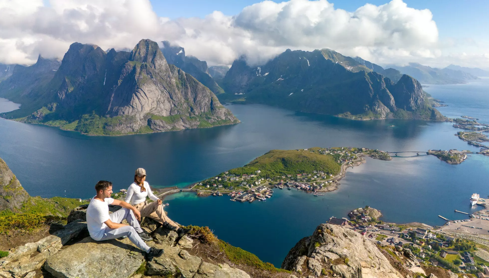 VAKKERT: Turgåere hviler på fjellkanten på Reinebringen i Lofoten, med utsikt over fjorder og øyer i solskinn. Illustrasjonsfoto: Fokke Baarssen / Shutterstock / NTB