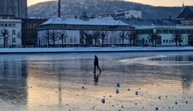 En enslig person går på den islagte overflaten av vannet, ikledd en svart jakke. Bakgrunnen viser bygninger, inkludert Bergen Kunsthall, omgitt av snøkledde trær. Sollyset reflekteres på isen, som har flere snøklumper spredt rundt. Bildet gir inntrykk av vinteraktiviteter i Bergen. (Bildebeskrivelsen er laget av en KI-tjeneste)