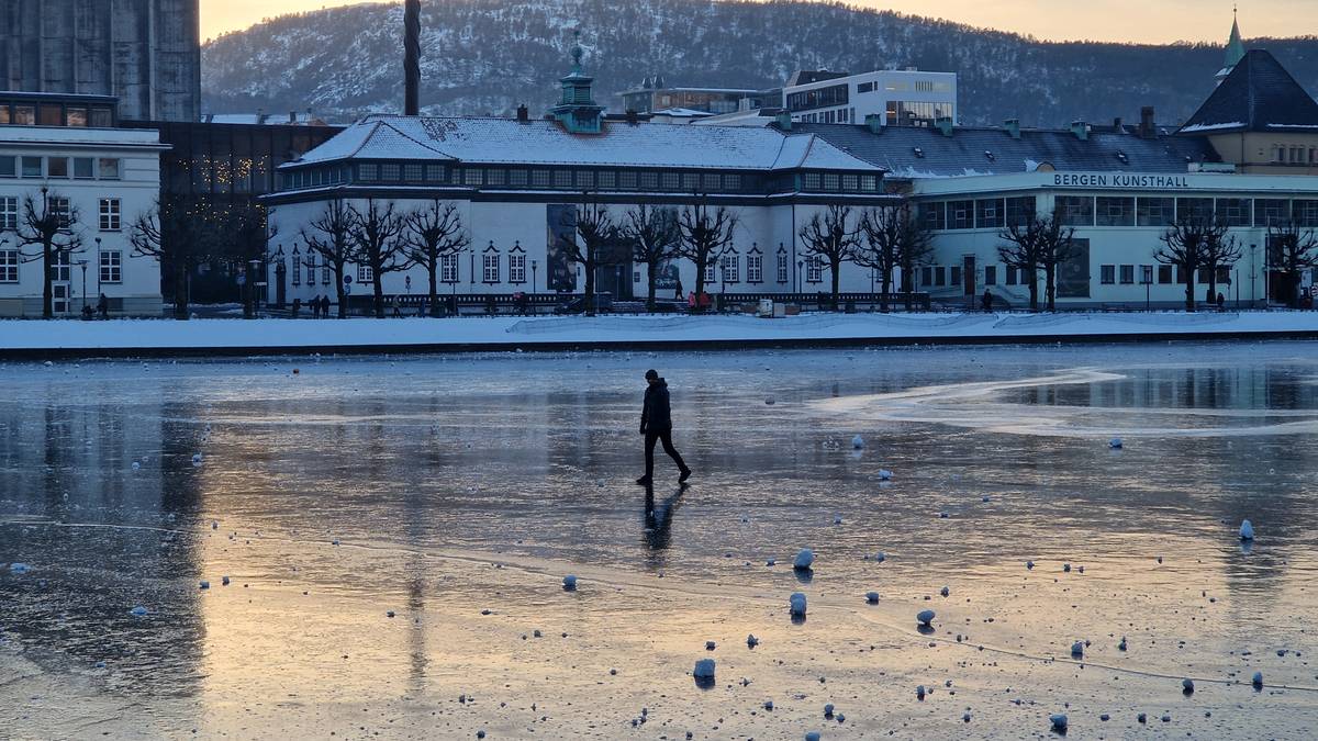 En enslig person går på den islagte overflaten av vannet, ikledd en svart jakke. Bakgrunnen viser bygninger, inkludert Bergen Kunsthall, omgitt av snøkledde trær. Sollyset reflekteres på isen, som har flere snøklumper spredt rundt. Bildet gir inntrykk av vinteraktiviteter i Bergen. (Bildebeskrivelsen er laget av en KI-tjeneste)