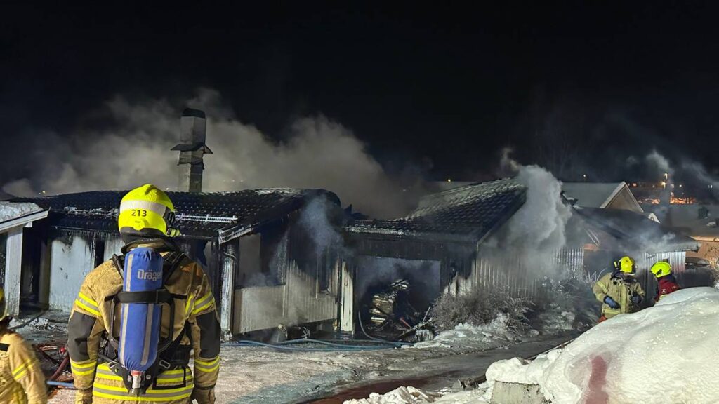 Brannmenn arbeider for å slukke en brann i en enebolig om natten. Røyk stiger fra de brente områdene av bygningen. Snø dekkes bakken rundt, og det er svake lys fra nabohusene i bakgrunnen. Brannmenn med hjelmer og brannvernutstyr er synlige i bildet. (Bildebeskrivelsen er laget av en KI-tjeneste)