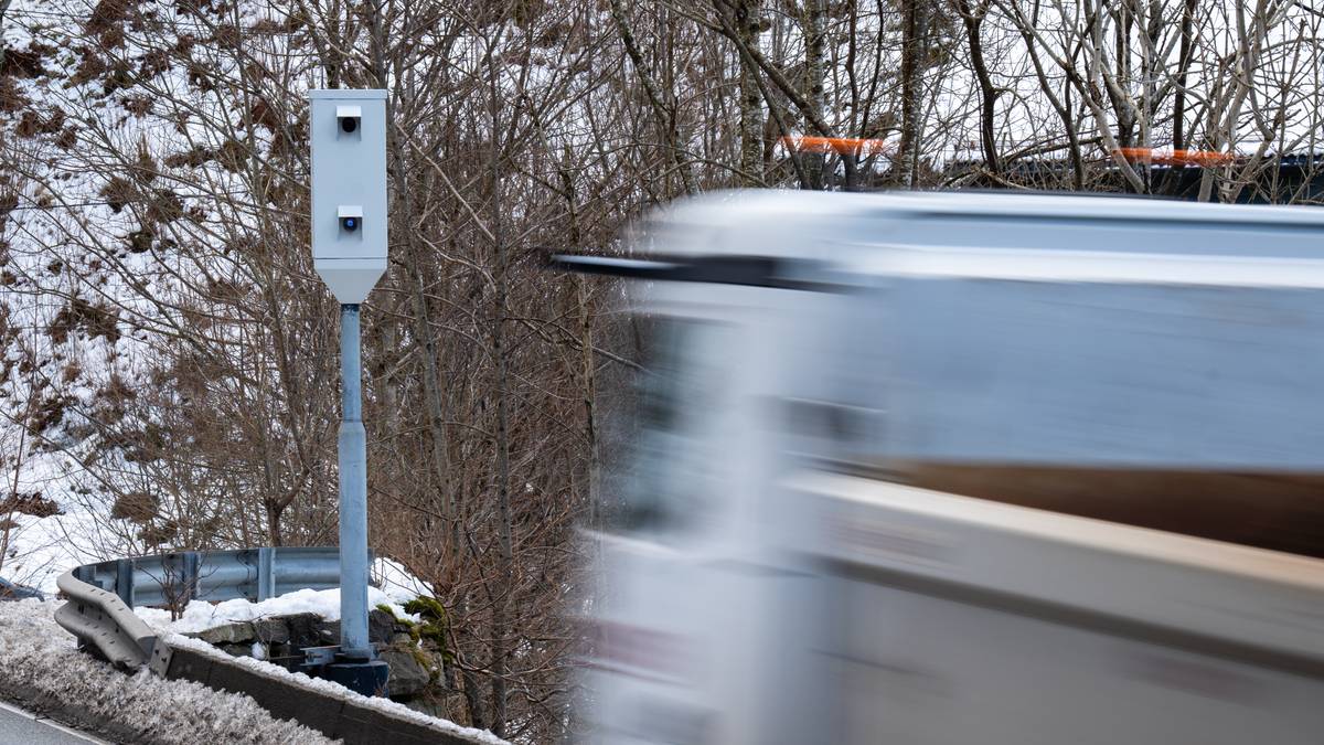 Et fartskamera er plassert langs veien, omgitt av snødekte bakkene. I forkant av kameraet passerer en lastebil i høy hastighet. Trær uten løv i bakgrunnen bidrar til et vinterlig landskap. Veikanten viser rester av snø og grus.