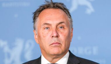 Nærbilde av Tore O. Sandvik som står foran en lyseblå vegg med Norges riksvåpen. Sandvik har grått hår og er iført en mørk dress.