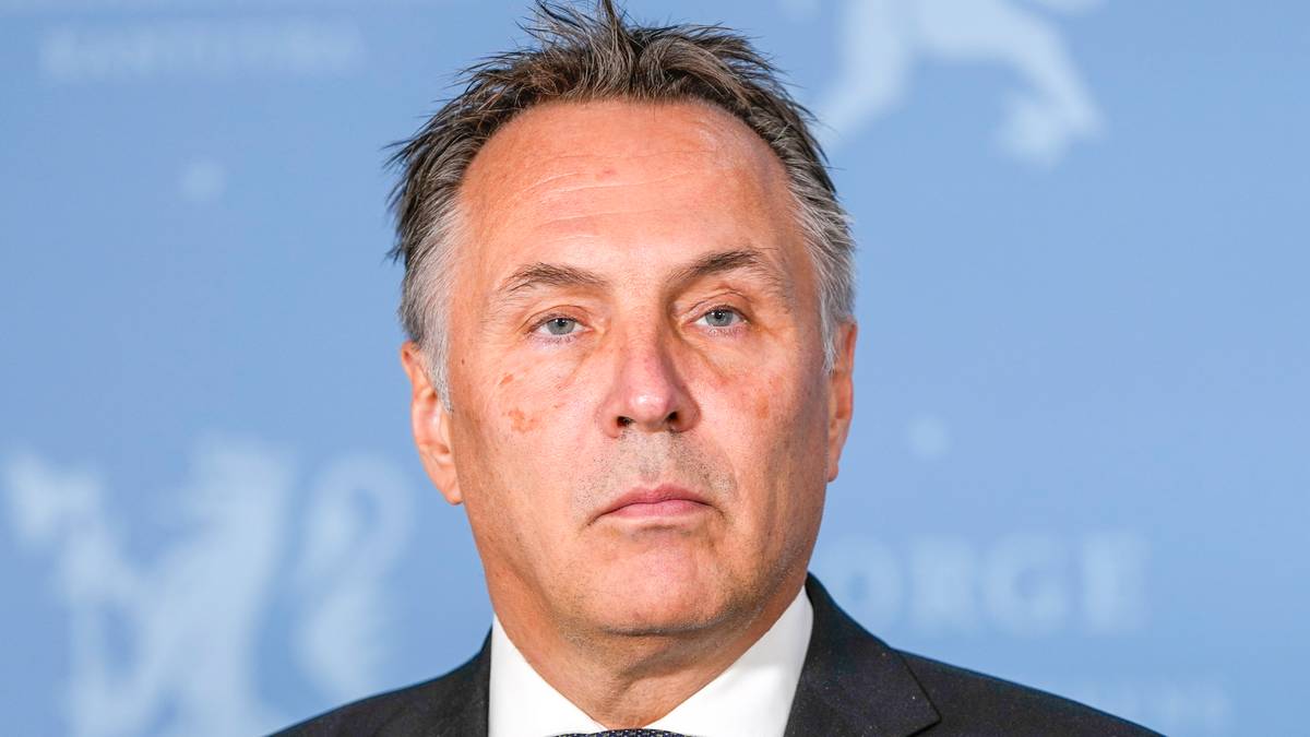 Nærbilde av Tore O. Sandvik som står foran en lyseblå vegg med Norges riksvåpen. Sandvik har grått hår og er iført en mørk dress.