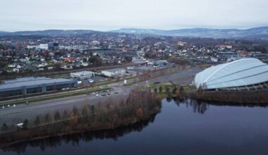 Bildet viser en oversiktsvisning av et urbant område med bygninger og veier i et grått vær. Til høyre synes en stor kuppelstruktur, sannsynligvis en idrettshall, som ligger nær en innsjø. Bakgrunnen viser et variert boligområde med hus i forskjellige stilarter og høyder. Området er preget av bilvei og parkeringsplasser, men det er få synlige mennesker.
