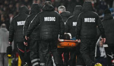 Flere medisinske fagfolk bærer en båre over en fotballbane. De er kledd i svarte jakker med "MEDIC" skrevet på ryggen. I bakgrunnen er delvis tilskuere synlige, og stadion er delvis dekket av regn. Båren synes å bære en person, men ansiktet er ikke synlig. (Bildebeskrivelsen er laget av en KI-tjeneste)