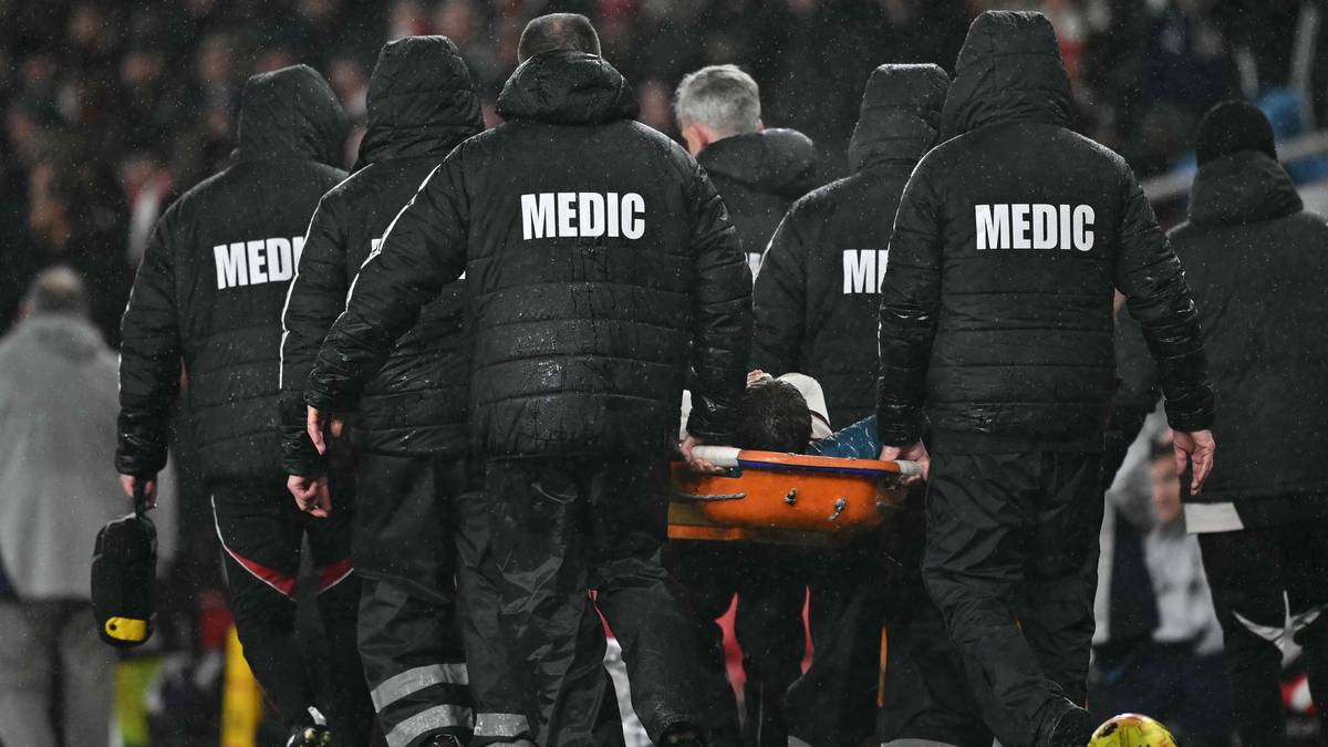Flere medisinske fagfolk bærer en båre over en fotballbane. De er kledd i svarte jakker med "MEDIC" skrevet på ryggen. I bakgrunnen er delvis tilskuere synlige, og stadion er delvis dekket av regn. Båren synes å bære en person, men ansiktet er ikke synlig. (Bildebeskrivelsen er laget av en KI-tjeneste)
