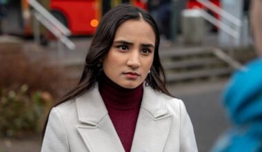 En kvinne med mørkt hår står fremfor kameraet iført en lys ytterjakke og rød genser. Bak henne ses en offentlig plass med trapp og busser i bakgrunnen. Det er en grå dag med lite folkeliv. Kvinnen ser mot en person til høyre utenfor synsfeltet.