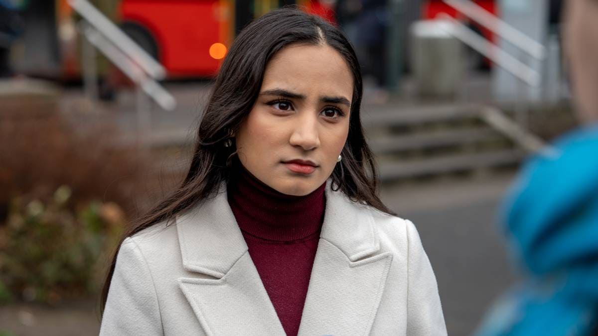 En kvinne med mørkt hår står fremfor kameraet iført en lys ytterjakke og rød genser. Bak henne ses en offentlig plass med trapp og busser i bakgrunnen. Det er en grå dag med lite folkeliv. Kvinnen ser mot en person til høyre utenfor synsfeltet.