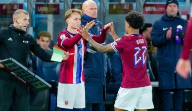 Bildet viser en fotballkamp der en spiller, iført en rød trøye med nummer 27, gjør seg klar til å byttes inn. Han hilser på en annen spiller, som har på seg en hvit trøye med nummer 10, før byttet skjer. En trener står med en digital bytteboks i hånden, og det er flere tilskuere i bakgrunnen. Omgivelsene er en idrettsarena med tydelig sportslig atmosfære. (Bildebeskrivelsen er laget av en KI-tjeneste)