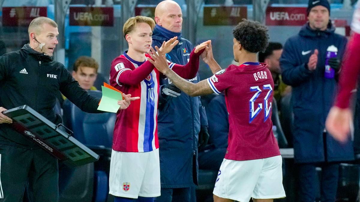 Bildet viser en fotballkamp der en spiller, iført en rød trøye med nummer 27, gjør seg klar til å byttes inn. Han hilser på en annen spiller, som har på seg en hvit trøye med nummer 10, før byttet skjer. En trener står med en digital bytteboks i hånden, og det er flere tilskuere i bakgrunnen. Omgivelsene er en idrettsarena med tydelig sportslig atmosfære. (Bildebeskrivelsen er laget av en KI-tjeneste)