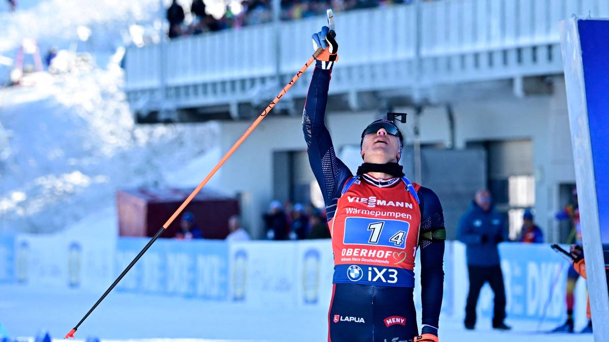 En skihopper feirer seieren etter et vellykket hopp, med staven hevet over hodet. Utøverens drakt er i lyse farger, med klærmerker synlige. I bakgrunnen kan man se en tilskuertribune og snødekte omgivelser. Arrangementet foregår sannsynligvis i en vinteridrettskontekst. (Bildebeskrivelsen er laget av en KI-tjeneste)