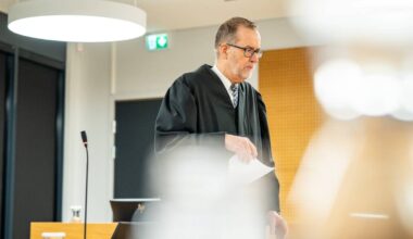 Statsadvokaten holder et papirark. Omgivelsene inkluderer moderne møbler og belysning. En mikrofon er synlig på bordet foran dommeren. Rettssalen ser ut til å være godt opplyst og organisert.