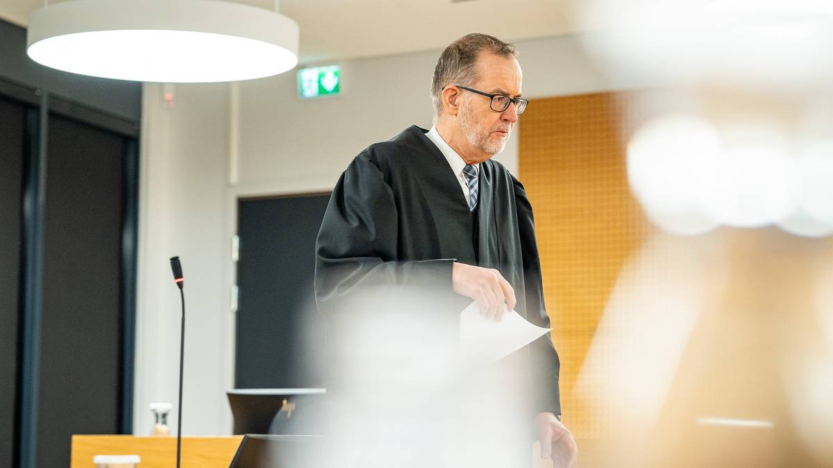 Statsadvokaten holder et papirark. Omgivelsene inkluderer moderne møbler og belysning. En mikrofon er synlig på bordet foran dommeren. Rettssalen ser ut til å være godt opplyst og organisert.