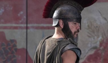 En person iført en romersk harnisk og hjelm med rød fjær står med ryggen til kameraet. Han har på seg en cappe og armbeskyttere, og ser ut til å være i en historisk setting med malerier i bakgrunnen. Omgivelsene inkluderer en veggdekorasjon som kan indikere et gammelt romersk tema. Bildet gir inntrykk av en dramatisk eller historisk fremstilling. (Bildebeskrivelsen er laget av en KI-tjeneste)