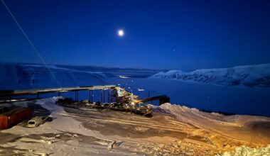 Bilde viser en snødekt landskap om natten, med månen svevende over horisonten. En abbannad struktur med lys er synlig, muligens en utpost eller forskningsstasjon nær en islagt sjø. Området er dominert av bratte fjell og snø, med spor av kjøretøy i snøen. Den klare nattehimmelen bidrar til en stille atmosfære. (Bildebeskrivelsen er laget av en KI-tjeneste)