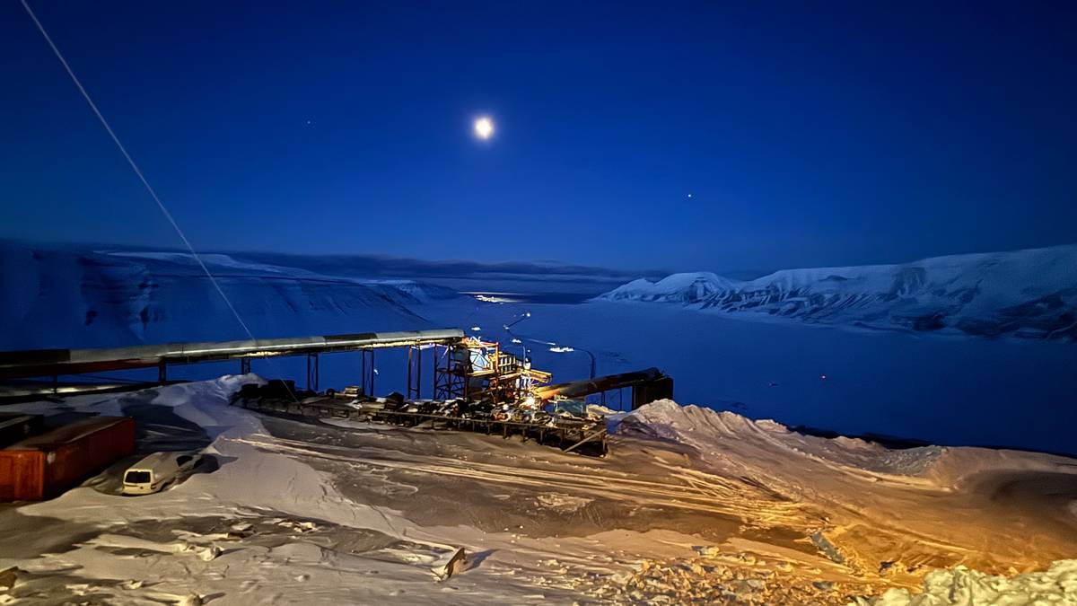 Bilde viser en snødekt landskap om natten, med månen svevende over horisonten. En abbannad struktur med lys er synlig, muligens en utpost eller forskningsstasjon nær en islagt sjø. Området er dominert av bratte fjell og snø, med spor av kjøretøy i snøen. Den klare nattehimmelen bidrar til en stille atmosfære. (Bildebeskrivelsen er laget av en KI-tjeneste)