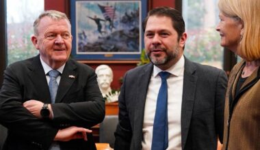 Tre personer står tett sammen i et rom. Den ene mannen har på seg en sort jakke med en skjorte og slips, mens den andre mannen er iført en mørk blazer og hvit skjorte med slips.