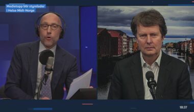 To personer deltar i en videokonferanse. Den ene personen sitter foran en mikrofon og holder et dokument, mens den andre snakker med et bylandskap i bakgrunnen. Begge er kledd i formelle klær. Mørkeblue bakgrunnsfarge dominerer venstre side av bildet.