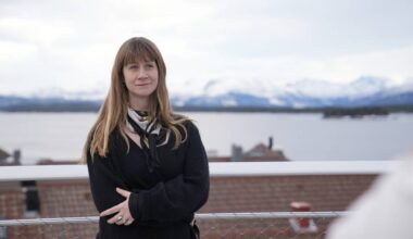 En person med langt, lyst hår står med armene kryssede foran seg. Vedkommende bærer en svart genser og et skjerf. Bakgrunnen viser et landskap med fjell og et vann, fylt med snødekte topper. Omgivelsene virker rolig, med hustak synlige i forkant.