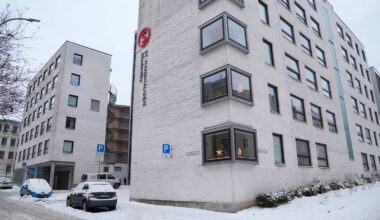 Bygningen av St. Hanshaugen, en moderne boligblokk, står i sentrum av bildet. Snøen dekker bakken og parkerte biler, mens en person er synlig til venstre, iført mørke klær. Det er flere vinduer på bygningen, noen med synlige dekorasjoner. Omgivelsene er preget av urbane elementer og en kald, vinterlig atmosfære. (Bildebeskrivelsen er laget av en KI-tjeneste)