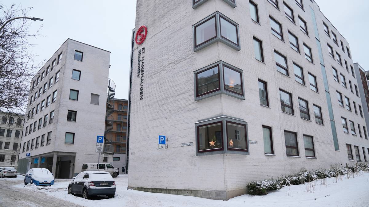 Bygningen av St. Hanshaugen, en moderne boligblokk, står i sentrum av bildet. Snøen dekker bakken og parkerte biler, mens en person er synlig til venstre, iført mørke klær. Det er flere vinduer på bygningen, noen med synlige dekorasjoner. Omgivelsene er preget av urbane elementer og en kald, vinterlig atmosfære. (Bildebeskrivelsen er laget av en KI-tjeneste)