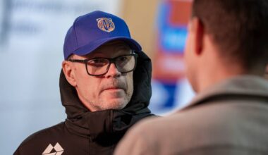 To personer er synlige i et intervju. Den ene har på seg en svart jakke med logo og blå caps, mens den andre har en lysere jakke. Omgivelsene er uklare, men det er flere bannere i bakgrunnen. Bildet fokuserer på samtalen mellom dem.