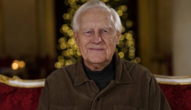 En eldre mann sitter på en rød stol med en dekorativ bakgrunn som har gyldne lys. Han har på seg en brun kordfløyelsjakke over en mørk genser. Omgivelsene virker festlige, noe som antyder en høytidelig anledning. Det er en dempet belysning i rommet, som bidrar til atmosfæren. (Bildebeskrivelsen er laget av en KI-tjeneste)