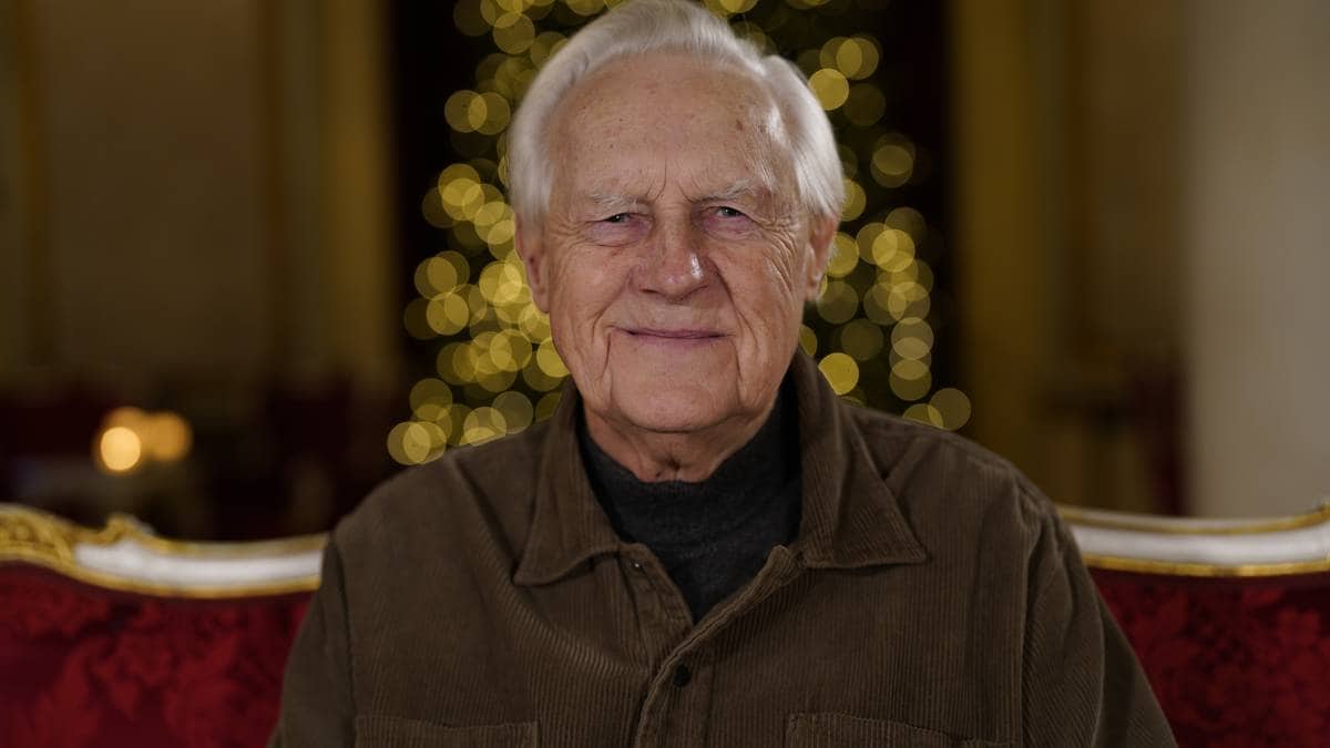 En eldre mann sitter på en rød stol med en dekorativ bakgrunn som har gyldne lys. Han har på seg en brun kordfløyelsjakke over en mørk genser. Omgivelsene virker festlige, noe som antyder en høytidelig anledning. Det er en dempet belysning i rommet, som bidrar til atmosfæren. (Bildebeskrivelsen er laget av en KI-tjeneste)