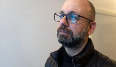 En voksen mann står ved en vegg i et rom med nøytral innredning. Han har på seg en svart genser og en svart, fyldig jakke. Mennesket har briller og kort, mørkt hår. Det er svakt lys i rommet, som skaper en rolig atmosfære.