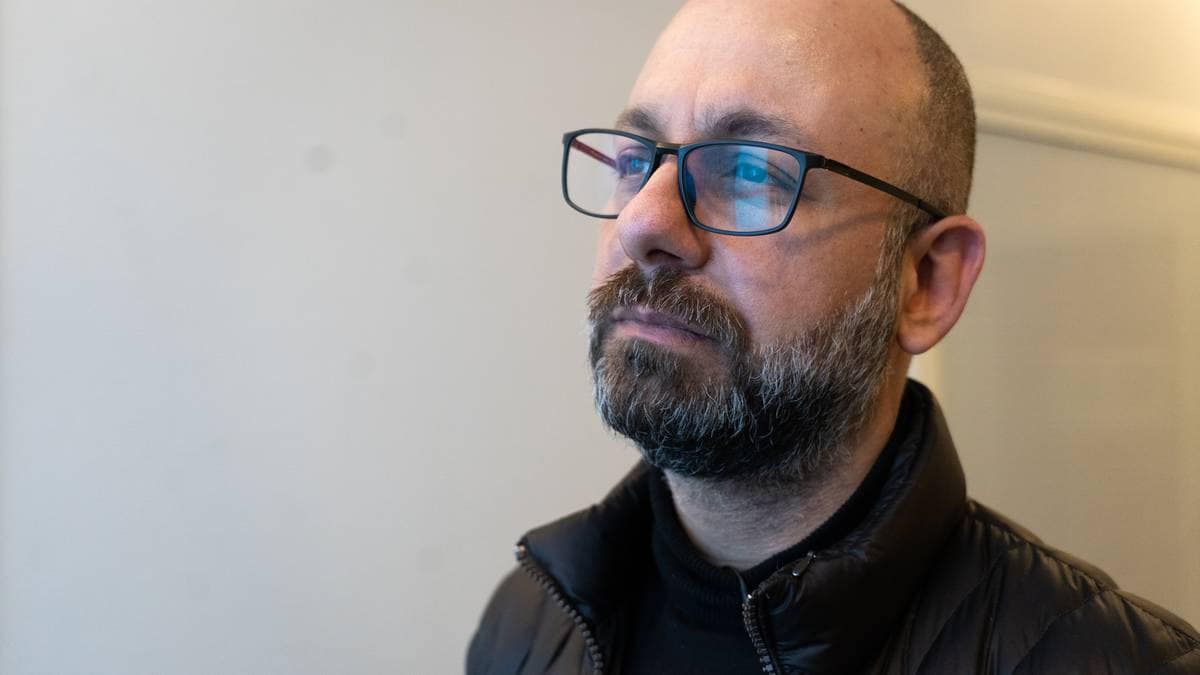 En voksen mann står ved en vegg i et rom med nøytral innredning. Han har på seg en svart genser og en svart, fyldig jakke. Mennesket har briller og kort, mørkt hår. Det er svakt lys i rommet, som skaper en rolig atmosfære.