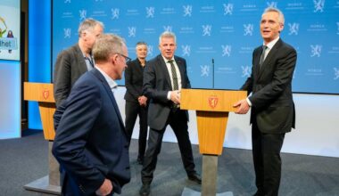 Fem personer står foran podiumer i en offisiell setting. Noen av dem er i samtale, mens en annen ser på. Det er skilting med offentlige symboler i bakgrunnen, og blå vegger danner en nøytral kontekst. Rommet har belysning som fremhever scenen, som virker tilrettelagt for en pressemelding. (Bildebeskrivelsen er laget av en KI-tjeneste)