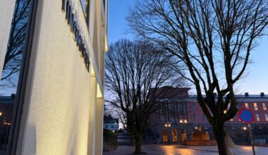 Bildet viser en gate med våte asfaltflater under morgenhimmelen. I forgrunnen er det en bygning med navnet "Tinghuset" på veggen, omgitt av nakne trær. Bakgrunnen viser flere bygninger med lys og en parkert bil ved veiskulderen. Lysene i området gir en dempet belysning i det tidlige morgenlyset. (Bildebeskrivelsen er laget av en KI-tjeneste)