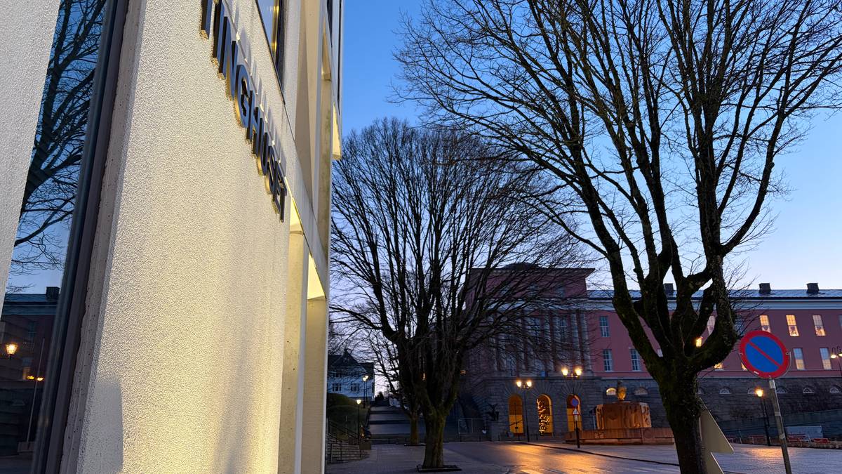 Bildet viser en gate med våte asfaltflater under morgenhimmelen. I forgrunnen er det en bygning med navnet "Tinghuset" på veggen, omgitt av nakne trær. Bakgrunnen viser flere bygninger med lys og en parkert bil ved veiskulderen. Lysene i området gir en dempet belysning i det tidlige morgenlyset. (Bildebeskrivelsen er laget av en KI-tjeneste)