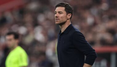 En fotballtrener står på sidelinja under en kamp, iført en mørk genser. Bakgrunnen viser en tilskuerstand med publikummere. En dommer i en gul drakt observerer spillet i nærheten. Stemningen er intens, typisk for en sportsbegivenhet. (Bildebeskrivelsen er laget av en KI-tjeneste)