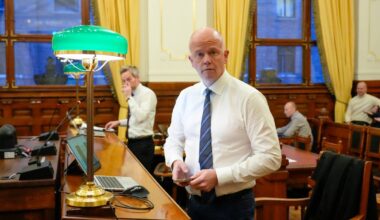 Bildet viser en person iført skjorte og slips, som står ved et skrivebord i et møterom. Bakgrunnen inkluderer store vinduer med gardiner og et grønnt lampe, samt flere personer i rommet. Noen er opptatt med å skrive eller se på skjermer, mens en annen person står til høyre. Interiøret gir et formelt preg, typisk for en offentlig samling. (Bildebeskrivelsen er laget av en KI-tjeneste)