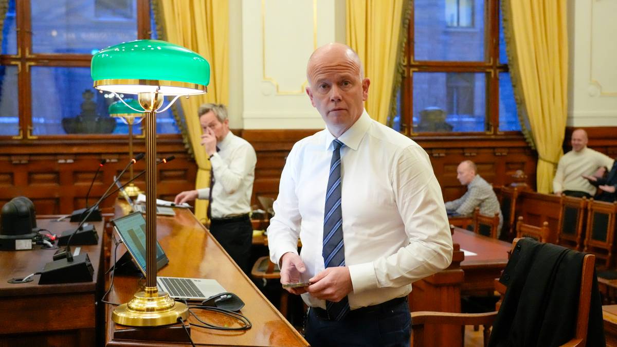 Bildet viser en person iført skjorte og slips, som står ved et skrivebord i et møterom. Bakgrunnen inkluderer store vinduer med gardiner og et grønnt lampe, samt flere personer i rommet. Noen er opptatt med å skrive eller se på skjermer, mens en annen person står til høyre. Interiøret gir et formelt preg, typisk for en offentlig samling. (Bildebeskrivelsen er laget av en KI-tjeneste)