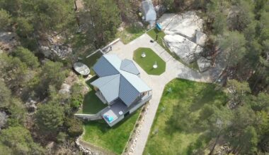 Bildet viser et moderne hus med grå tak i et skogkledd område. Omsluttende grønne plener og stier er synlige rundt bygningen. Det er noen mennesker i nærheten av huset som arbeider, samt steiner og trær i omgivelsene. Huset er plassert i et naturskjønt landskap. (Bildebeskrivelsen er laget av en KI-tjeneste)