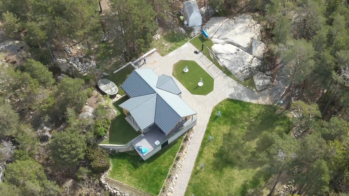 Bildet viser et moderne hus med grå tak i et skogkledd område. Omsluttende grønne plener og stier er synlige rundt bygningen. Det er noen mennesker i nærheten av huset som arbeider, samt steiner og trær i omgivelsene. Huset er plassert i et naturskjønt landskap. (Bildebeskrivelsen er laget av en KI-tjeneste)