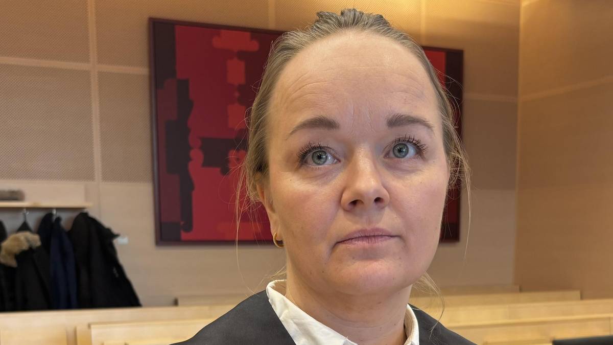 En kvinne i rettsdrakt står i en rettssal. Hun har blont hår og har på seg en hvit skjorte under drakten. Bak henne er flere bambus-stoler og en eller flere jakker hengende på en kleshenger. Veggen bak er dekorert med et kunstverk i sterke farger.