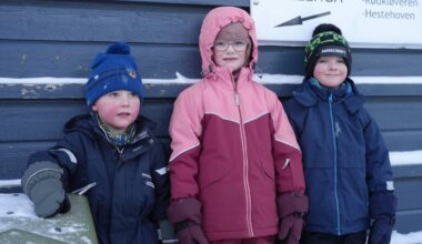 Tre barn står sammen foran en bygning. De er iført fargerike vinterklær, inkludert luer og hansker. Bakgrunnen viser en mørk vegg med snø og et skilt som indikert retningen til "Lilleenga". Omgivelsene tyder på vinter og kalde forhold. (Bildebeskrivelsen er laget av en KI-tjeneste)