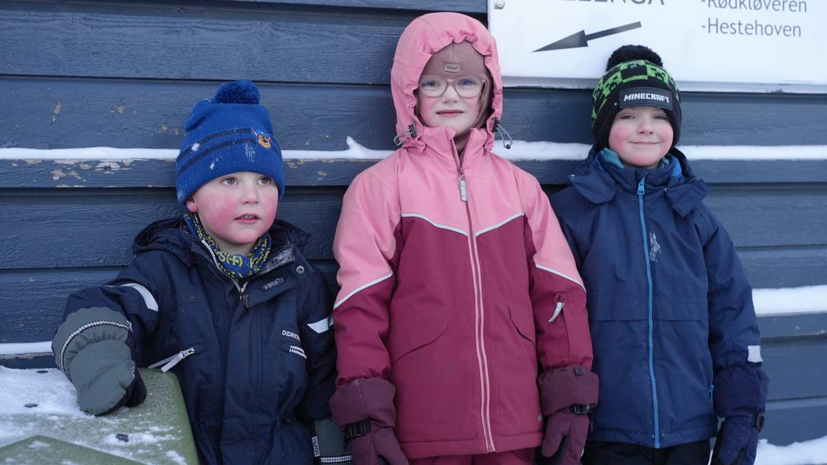 Tre barn står sammen foran en bygning. De er iført fargerike vinterklær, inkludert luer og hansker. Bakgrunnen viser en mørk vegg med snø og et skilt som indikert retningen til "Lilleenga". Omgivelsene tyder på vinter og kalde forhold. (Bildebeskrivelsen er laget av en KI-tjeneste)