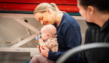 En kvinne holder et lite barn på fanget, som holder en leke med mange farger. Begge ser ut til å være i et rom med medisinsk utstyr i bakgrunnen. En annen person i mørk genser sitter i nærheten, men er delvis ute av fokus. Omgivelsene virker rolige, og opplever en uformell situasjon. (Bildebeskrivelsen er laget av en KI-tjeneste)