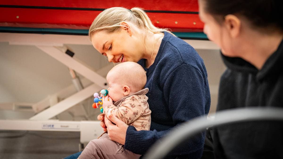 En kvinne holder et lite barn på fanget, som holder en leke med mange farger. Begge ser ut til å være i et rom med medisinsk utstyr i bakgrunnen. En annen person i mørk genser sitter i nærheten, men er delvis ute av fokus. Omgivelsene virker rolige, og opplever en uformell situasjon. (Bildebeskrivelsen er laget av en KI-tjeneste)
