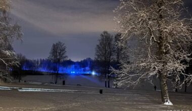 Bildet viser et snødekt landskap om natten, opplyst av blålys fra en eller flere kjøretøy i bakgrunnen. Trær med snø på greinene rammer inn scenen, og det er spor i snøen som indikerer aktivitet. I midten av bildet er det en søppelbøtte og benker. Bakgrunnen viser en vannoverflate med silhuetter av trær og bygninger. (Bildebeskrivelsen er laget av en KI-tjeneste)