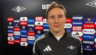 Et medlem av Rosenborg Ballklub står foran et veggbanner med sponsorer, i en sportslig atmosfære. Personen er kledd i en svart RBK-jakke med logoer. Bakgrunnen inneholder flere logoer fra sponsorer og arrangementer. Bildeutsnittet fokuserer på personen, som gir en profesjonell setting, typisk for pressevent. (Bildebeskrivelsen er laget av en KI-tjeneste)